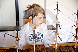 Little blond school kid boy playing with sailing ship model indoors. Excited child with yacht having fun after school at