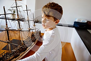 Little blond school kid boy playing with sailing ship model indoors. Excited child with yacht having fun after school at