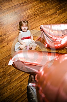 Little blonde baby girl two years old with big pink and white balloons lying on the wooden floor on her birthday party