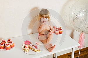 Little blonde baby girl two years old in pink pants sitting on the white table near her birthday cake and different pink sweets on