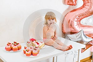 Little blonde baby girl two years old in pink pants sitting on the white table near her birthday cake and different pink sweets on