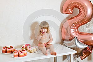 Little blonde baby girl two years old in pink pants sitting on the white table near her birthday cake and different pink sweets on