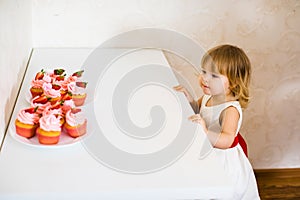 Little blonde baby girl two years old in white dress looking at her birthday cake and different pin sweets on the table