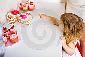 Little blonde baby girl two years old in white dress looking at her birthday cake and different pin sweets on the table