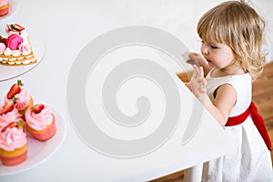 Little blonde baby girl two years old in white dress looking at her birthday cake and different pin sweets on the table