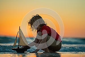 Little blonde boy put toy boat in the sea waves at the beach during summer vacation. Childhood and summer family