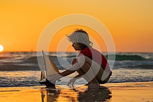 Little blonde boy put toy boat in the sea waves at the beach during summer vacation. Childhood and summer family