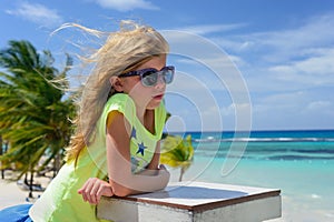 Little blonde caucasian girl in sunglasses is on the beach. Blue sky, ocean and palms are as a background.