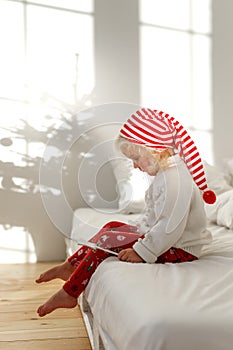 Little blonde child in Christmas hat sits on bed with white bedclothes, plays with magic wand, has serious concentrated