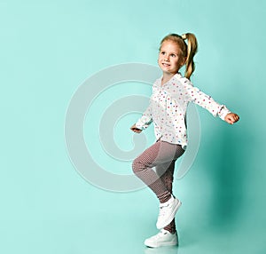 Little blonde child in shirt with hearts print, checkered pants, white sneakers. Smiling, posing on blue background. Full length