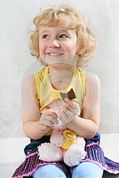 Little blonde curly girl eating chocolate with toy