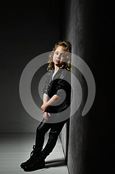 Little blonde curly kid in black leather jacket, pants and boots. She is standing leaning on gray wall in twilight of dark studio