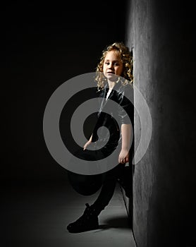 Little blonde curly kid in black leather jacket, pants and boots. She is standing leaning on gray wall in twilight of dark studio