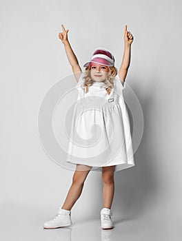 Little blonde curly positive princess girl in white casual dress and sneakers standing dancing walking and smiling  over grey wall