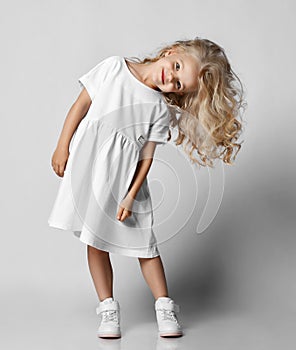 Little blonde curly positive princess girl in white casual dress and sneakers standing and playing with hair