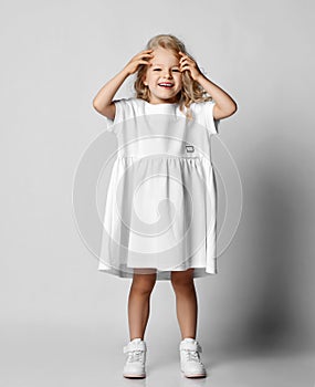 Little blonde curly positive princess girl in white casual dress and sneakers standing, touching hair and smiling