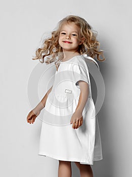 Little blonde curly positive princess girl in white casual dress and sneakers standing walking with curly hair over grey wall