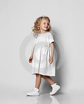 Little blonde curly positive princess girl in white casual dress and sneakers standing walking with curly hair over grey wall