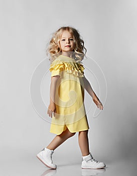 Little blonde curly positive princess girl in white casual dress and sneakers standing walking with curly hair over grey wall
