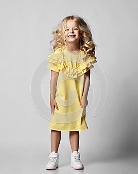 Little blonde curly positive princess girl in white casual dress and sneakers standing walking with curly hair over grey wall