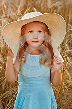 A little blonde girl in a blue dress and a very big mother`s hat on a wheat field at sunset. Portrait of a child against the