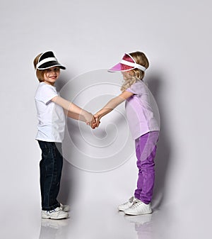 Little blonde girl and boy in colorful casual clothes and sun visors. Smiling, looking at each other, posing isolated on white