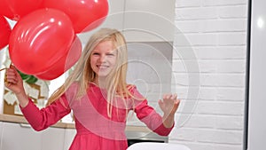Little blonde girl dancing with red heart shape balloons at home. Celebrating Valentines Day. Slow motion.