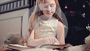 A little blonde girl in a festive dress with a book in her hands sits next to soft toys against the background of a