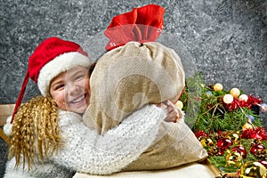 Little blonde girl in a gnome`s red hat with santa`s bag in anticipation of a Christmas miracle hugs a bag with Christmas gifts