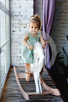 A little blonde girl is having fun and swinging on a wooden rocking horse near the window