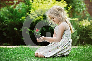 Little blonde girl holding young flower plant in hands on green background