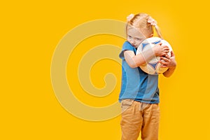 Little blonde girl hugs soccer ball. yellow studio background. Copy space, mockup. Football for kids