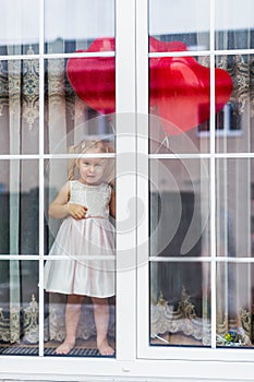 Little blonde girl in a light dress with heart-shaped baloons staying nead a big window at home. Shoot from outside