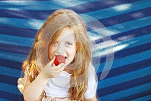 little blonde girl with long hair eats a strawberry in the garden sitting in a blue hammock
