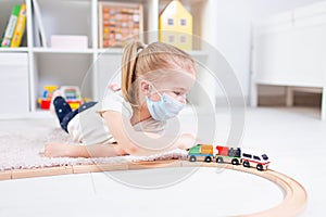 Little blonde girl in a medical mask playing with toy train on a floor in a light room at hone during quarantine