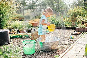 Little blonde girl playing at garden with water in a tin basin. Kids gardening. Summer outdoor water fun. Childhood in the country