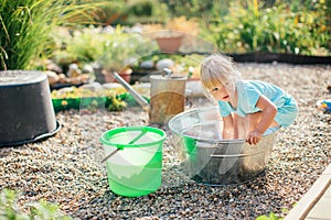 Little blonde girl playing at garden with water in a tin basin. Kids gardening. Summer outdoor water fun. Childhood in the country