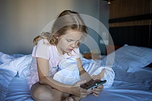 A little blonde girl is playing with her smartphone while sitting in a snow-white bed. The harm of gadgets for children