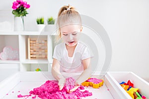 Little blonde girl playing with pink kinetic sand on a white table in a light room. Sensory development. Lessons in a kindergarten