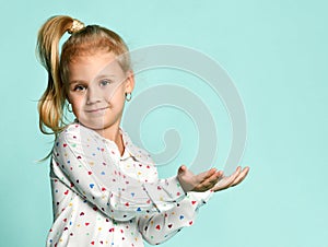 Little blonde girl with ponytail, in shirt with hearts print. She smiling, holding something posing on blue background. Close up