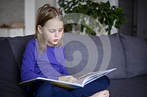 a little blonde girl is reading a big book sitting on the sofa in the living room.Smile and interest in reading.Self