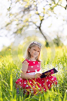 Little blonde girl in a red dress reads a book with fairy tales in nature