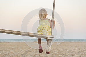 Little blonde girl sits on the edge of wooden bench-swing sea background and watches the sunset. Yellow magic sunset lights
