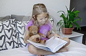 A little blonde girl is sitting on the bed before going to bed, hugging her teddy bear and reading him a children's