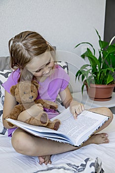 A little blonde girl is sitting on the bed before going to bed, hugging her teddy bear and reading him a children& x27;s