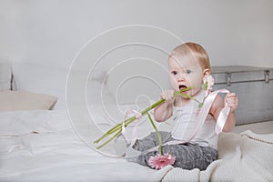Little blonde girl sitting on a bed with pink flowers and a ribbon and biting them, portrait of a cute baby on a light background