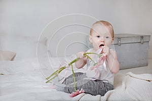 Blonde girl sitting on a bed with pink flowers and ribbon, portrait of a cute baby on a light background