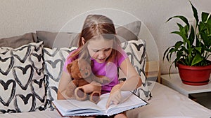 A little blonde girl is sitting in her bed on a white sheet before going to bed, hugging a teddy bear and reading a