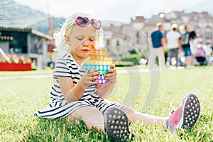 A little blonde girl sitting on the lawn plays with a trendy toy pop it.