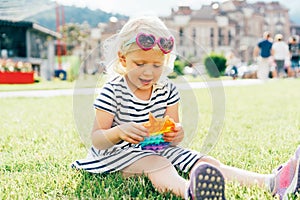 A little blonde girl sitting on the lawn plays with a trendy toy pop it.